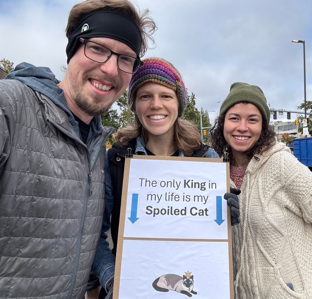 Three protestors hold a sign that says “The only KING in my life is my Spoiled Cat” followed by a drawing of a handsome cat wearing a crown 