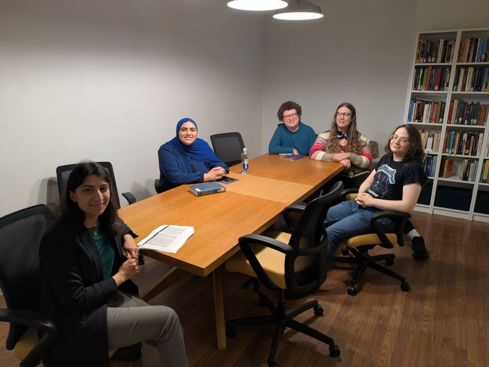 Four students around a conference table with Lina Khan on the other end. There is a bookshelf behind the students. 