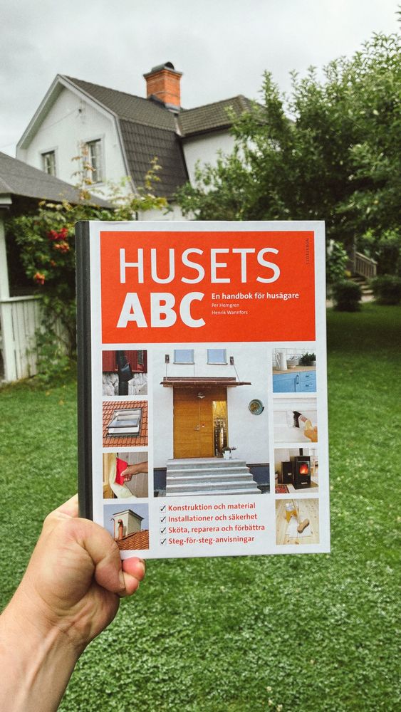I förgrunden håller en hand håller upp boken ”Husets ABC”. I bakgrunden syns en grönskande trädgård och ett vitt hus.