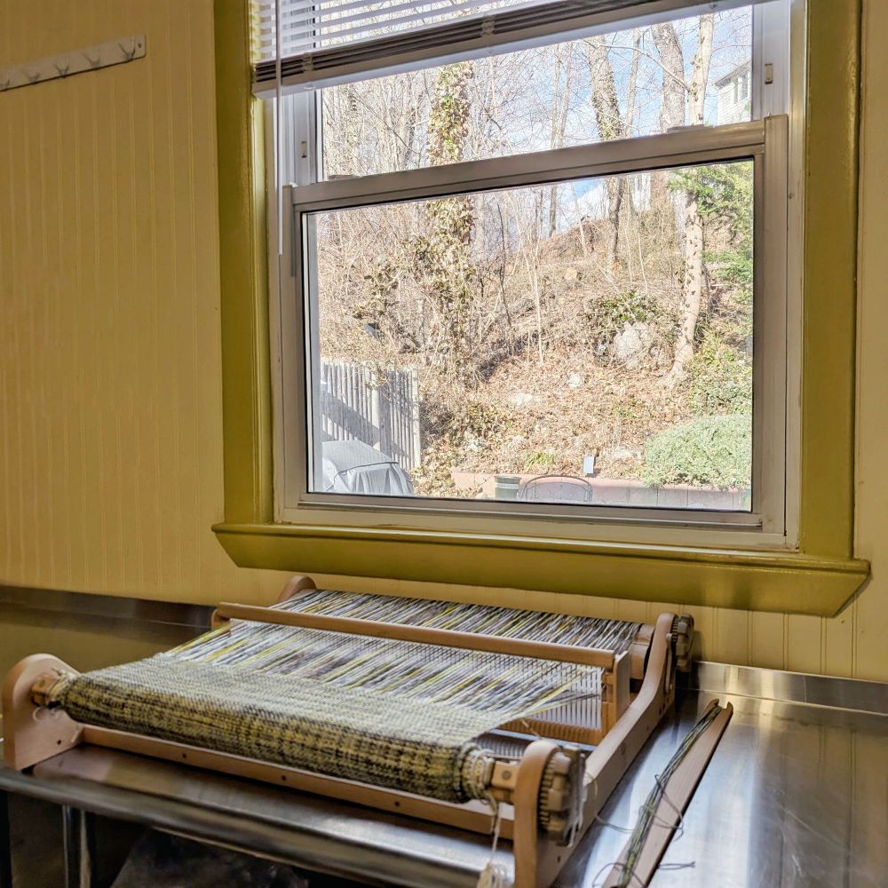 A rigid heddle loom sits on a stainless steel work table against a yellow wall facing a window. Outside the window is a hill with trees, bare from the winter season, and the corner of a wooden fence.