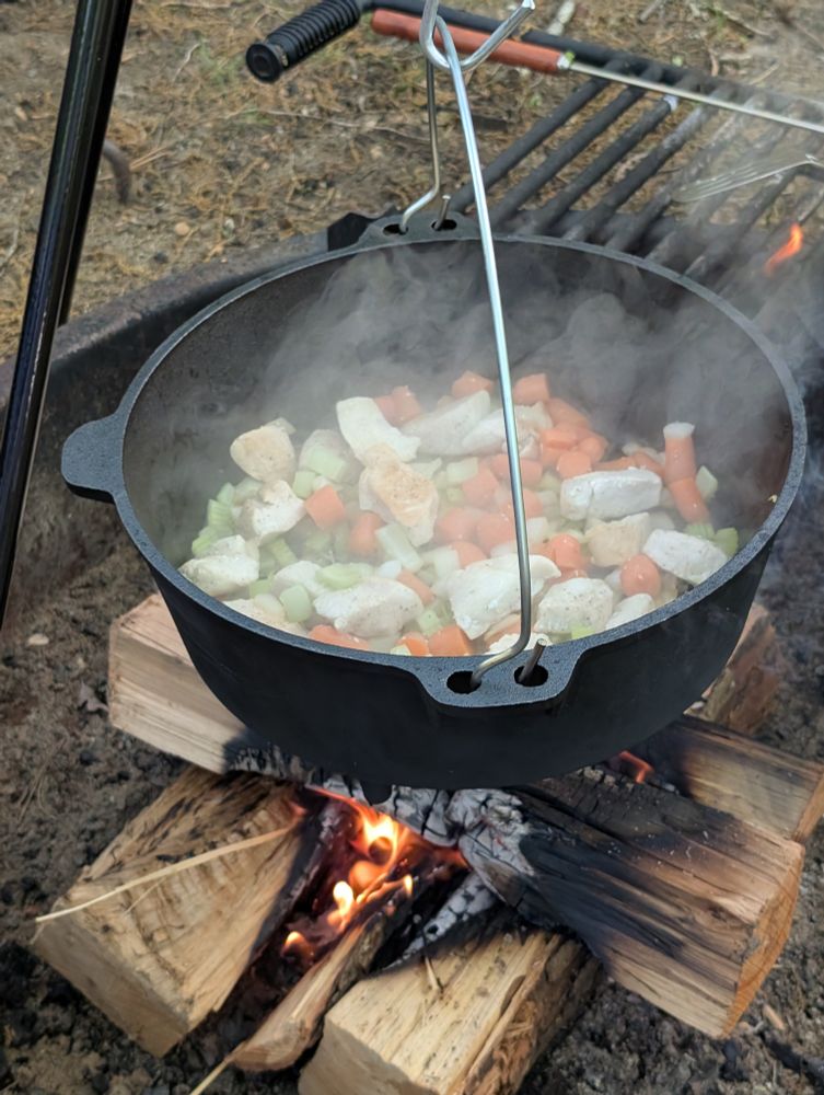 Chicken and vegetables cook in an open pot hanging over a wood campfire.