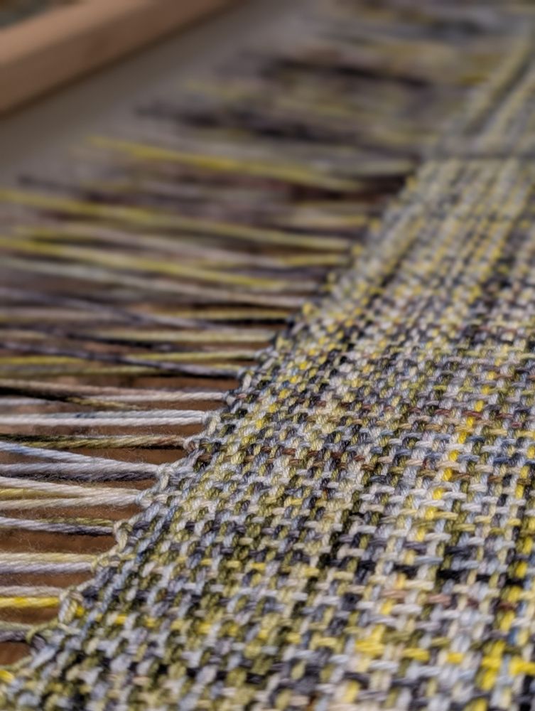 A close-up of handwoven fabric still on the loom. The left side shows the remaining warp threads with hand stitched knots against the finished fabric to the right. The coloring is variegated with yellow, pale blue, green, and almost black.