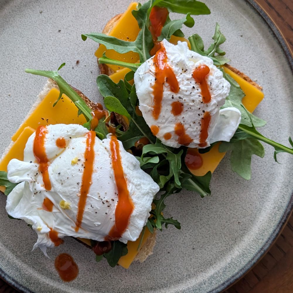 Viewed from above, two poached eggs on a bed of arugula, cheddar cheese, and lightly toasted homemade sourdough bread. Salt, pepper, and Sriracha sauce top the eggs.