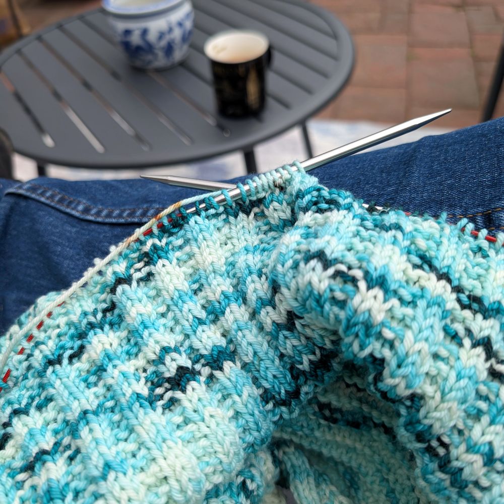 A blue and white variegated knitting project on metal circular cable knitting needles. In the background there is a round outdoor table with a coffee mug and a blue and white pot in the center.