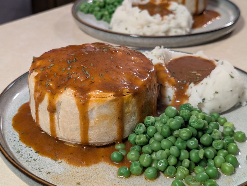 A pie played with gravy dripping down the sides, next to mashed potatoes and peas.