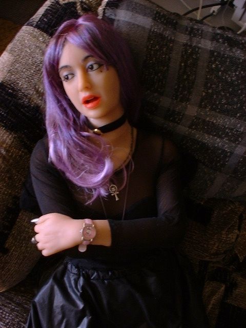 A pale-skinned companion Doll with long lavender hair, a black velveteen choker with a purple jewel in the centre, and a black cotton and mesh top, reclines on a grey loveseat, looking off to her right, Her rubbery red lips are parted and her arms are folded across her chest.