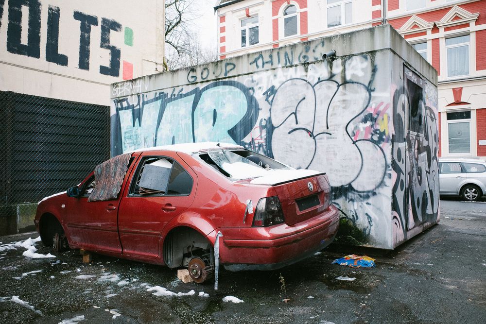 Ein Foto von einem komplett ramponierten Auto vor einer mit Graffiti besprühten Wand. Das Auto hat keine Reifen mehr, sondern ist auf Ziegelsteinen aufgebockt. Alle Scheiben sind kaputt, ein paar Fetzen Klebeband hängen an der Seite, das Nummernschild fehlt.