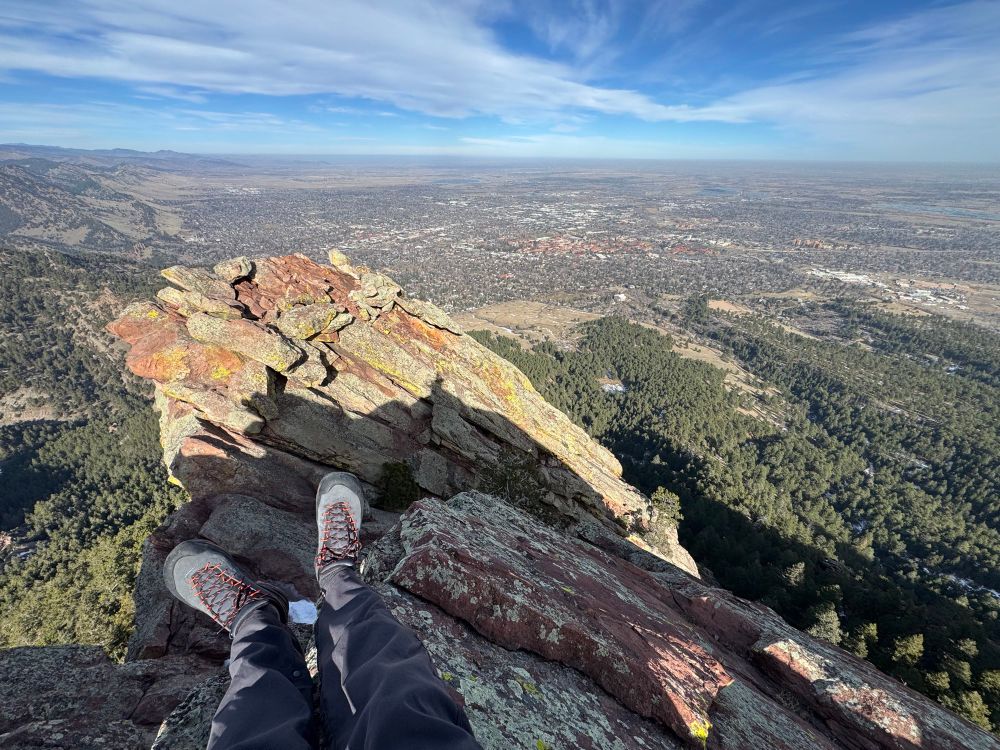 Legs and feet in the foreground of a person sitting on a rock. The frame is a high up view of Boulder, CO from the top of the first flatiron. 