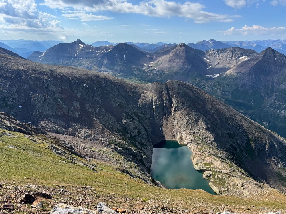 View looking down at an alpine lake with wispy clouds and mountain peaks in the background
