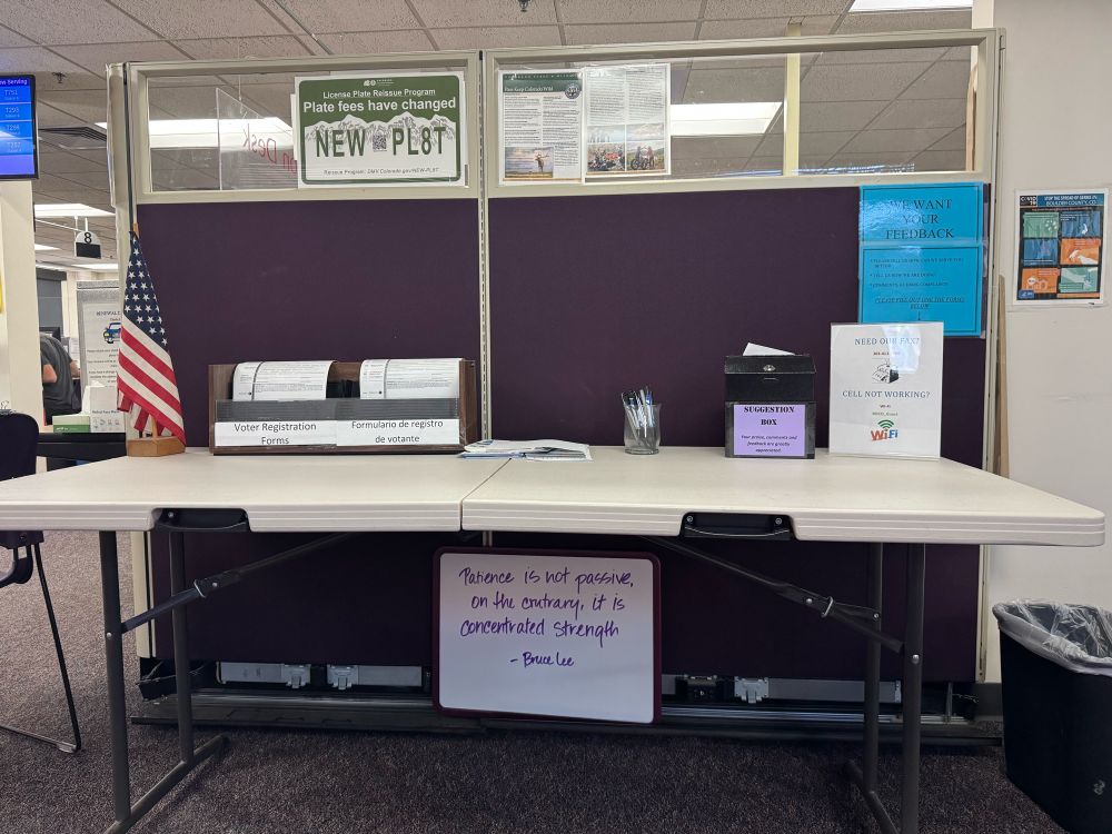 A whiteboard, under a folding table with an American flag and a cubicle wall with a DMV vibe, hand written quoting “patience is not passive, on the contrary, it is concentrated strength” - Bruce Lee