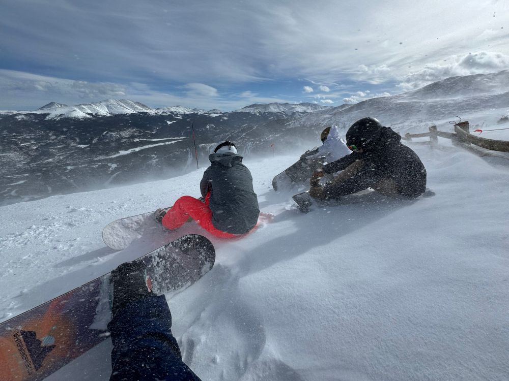First person view of a snowboarder with three other boarders in view, strapping in for a run. There’s blowing snow in frame and a snow fence, with a mountain vista in the background.