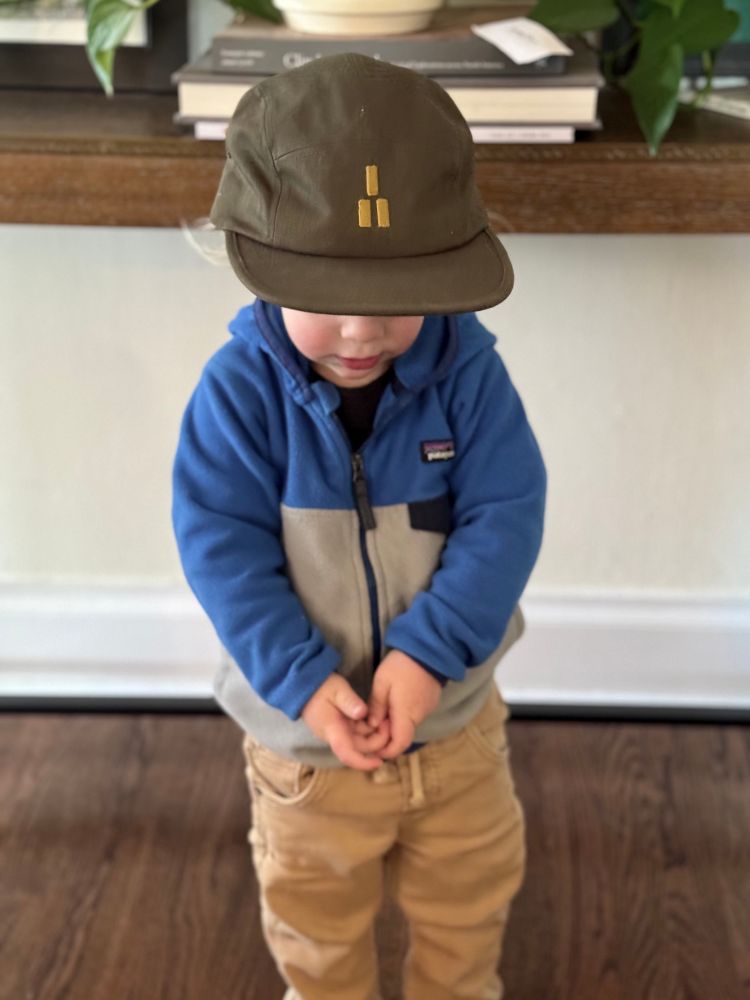 A toddler wearing a blue and gray zip up fleece and brown pants has a hat with the Trails Magazine blaze logo on it, just obscuring his face. It’s way too big to fit his head but no matter. 