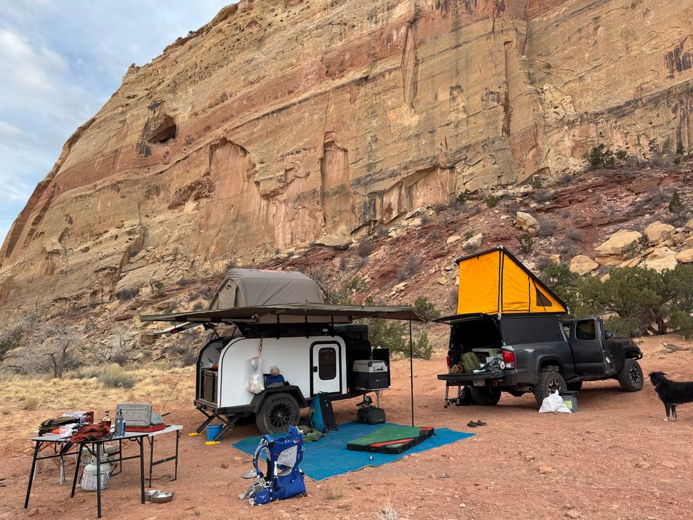 Dave’s third gen Tacoma with a GoFast camper popped up, with an all terrain camper trailer behind it. The trailer has an awning and tent deployed, and a blue mat in front of it. A very good dog is just in frame right.