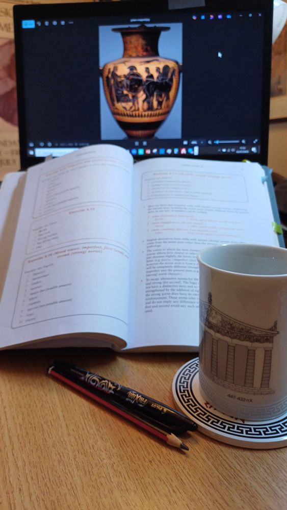 An open laptop displaying an image of an ancient Greek vase, an open textbook, pen and pencil, and a mug with a picture of the Parthenon  on it.
