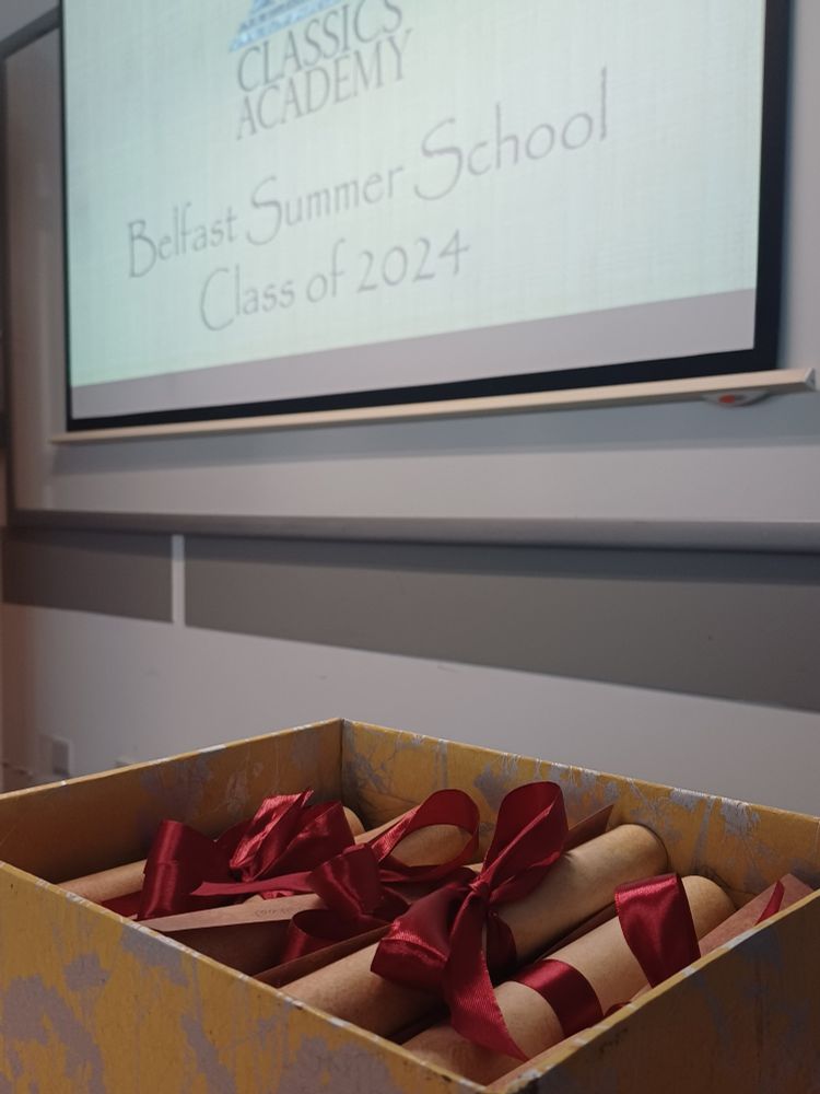 Projector screen reading "H.M. Classics Academy, Belfast Summer School, Class of 2024" and a box containing scrolls tied with red ribbon