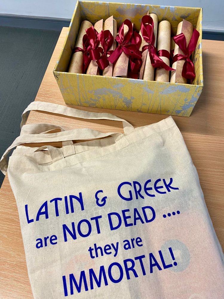 A cream coloured tote bag bearing the words "Latin and Greek are not dead ... they are immortal" next to a box containing scrolls tied with red ribbon 