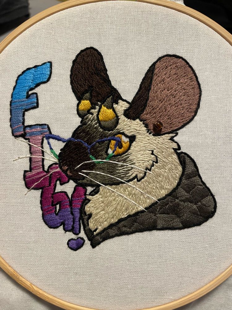 An embroidered tan, seal point mouse with a yellow eye, horns, wearing glasses,  looking at the viewer. It wears a hood around its neck, where the art ends. There is a single bead acting as an earring. The text, vertical, reads FIG!