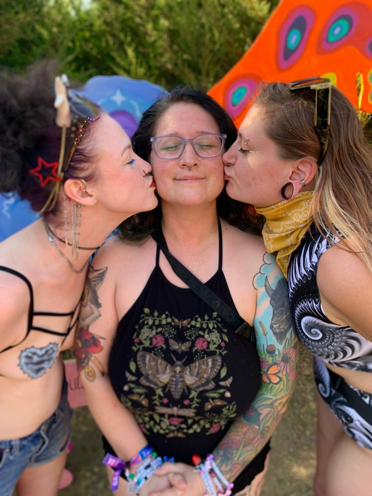 Three girls stand together. The two on the outside lean in to kiss the girl in the middle. They are at Symmetry Music Festival 2025.