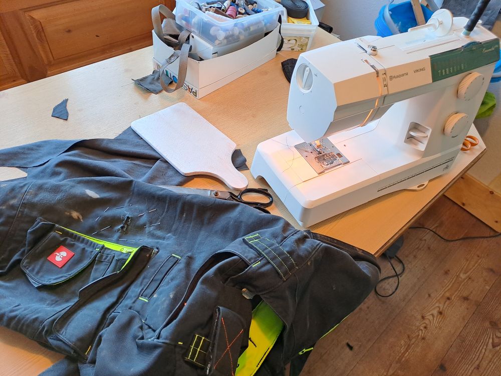 Eine Nähmaschine auf einem Tisch, daneben verschiedene Utensilien, die gebraucht werden. Links von der Nähmaschine eine schwarze Arbeits-Latzhose.