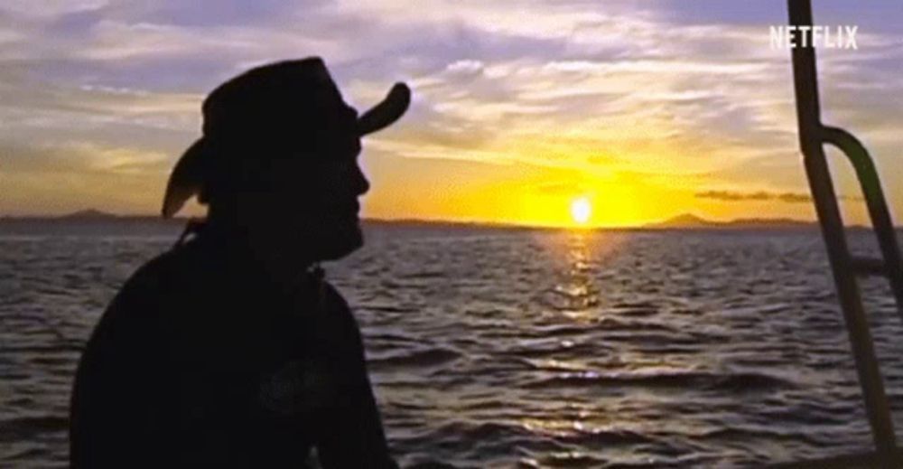 a man in a cowboy hat is sitting on a boat watching the sun set behind him