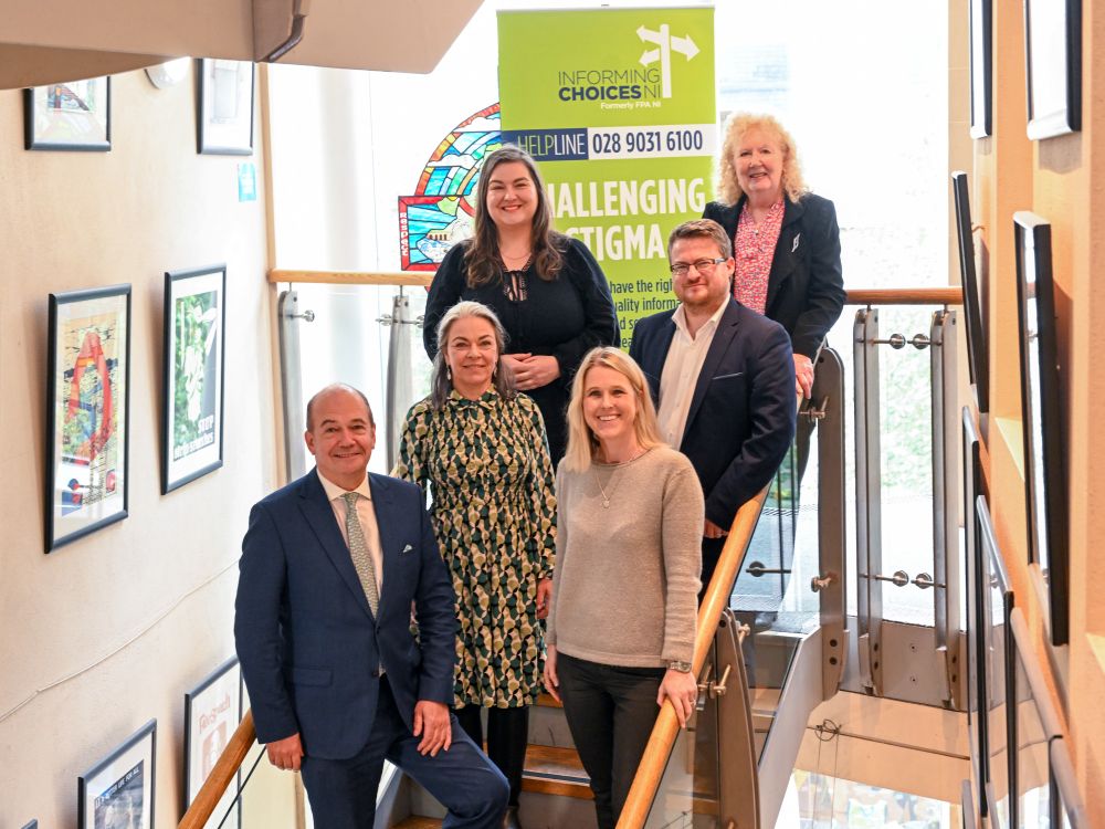 The Informing Choices NI team are stood on a staircase with a green poster behind them that says 'Challenging Stigma'. The team are smiling at the camera. 