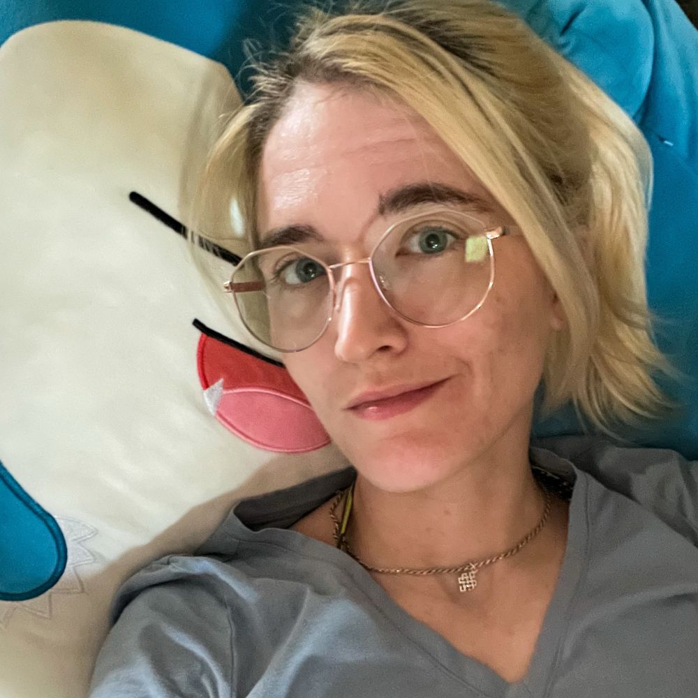 Selfie showing off new glasses. Hair is currently blonde in a ponytail and I’m laying back on a large Snorlax squishmallow. 