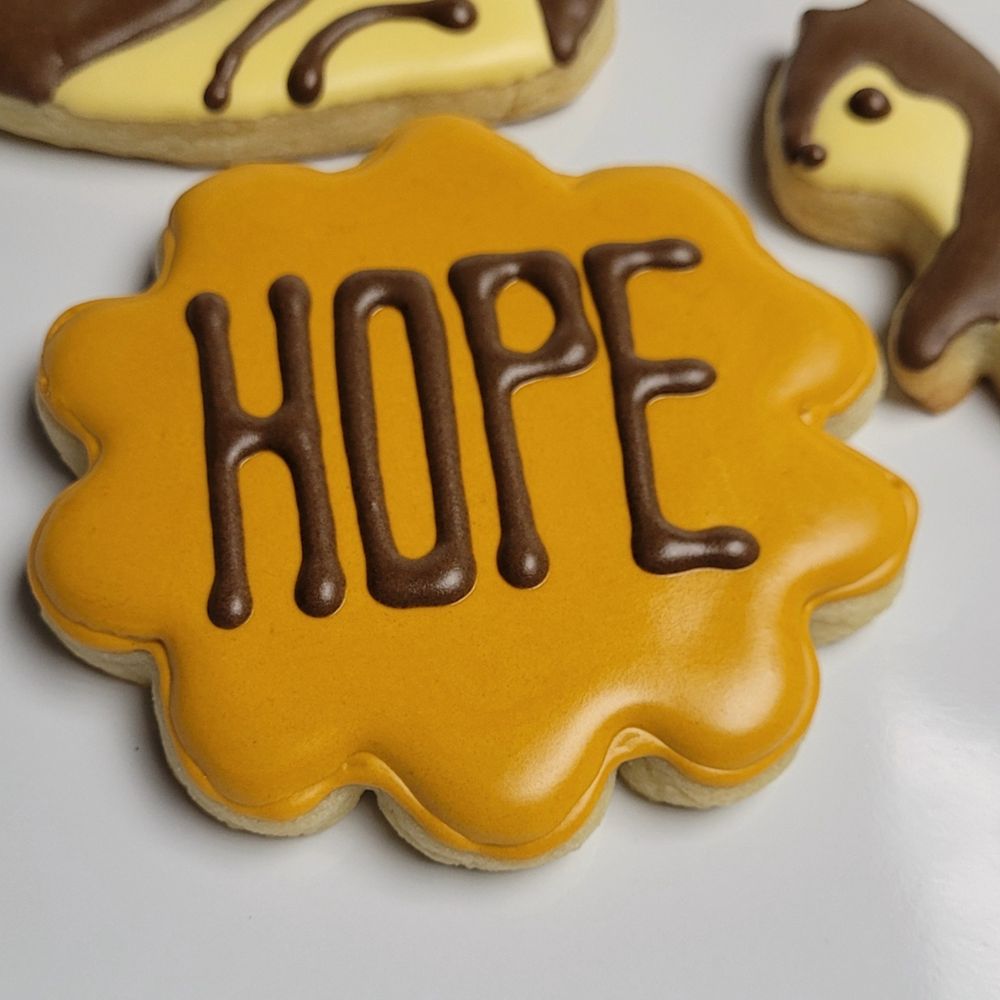 a flower cookie flooded with golden yellow royal icing with the word "HOPE" written in brown. cookie is on a white background with parts of brown squirrels showing.