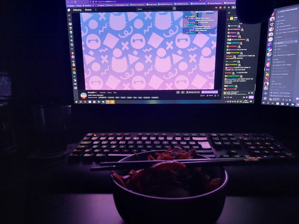 A bowl of dumplings and a veggie wok with two metal chopsticks on top of it. SocksBX's stream is on in the background