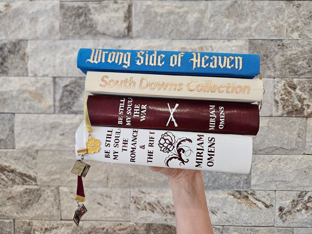 A pile of hand bound hardcover books with texts "Wrong Side Of Heaven", "South Downs Collection", "Be Still My Soul: The War", "Be Still My Soul: The Romance & The Rift", all written by Mirjam Omens. 