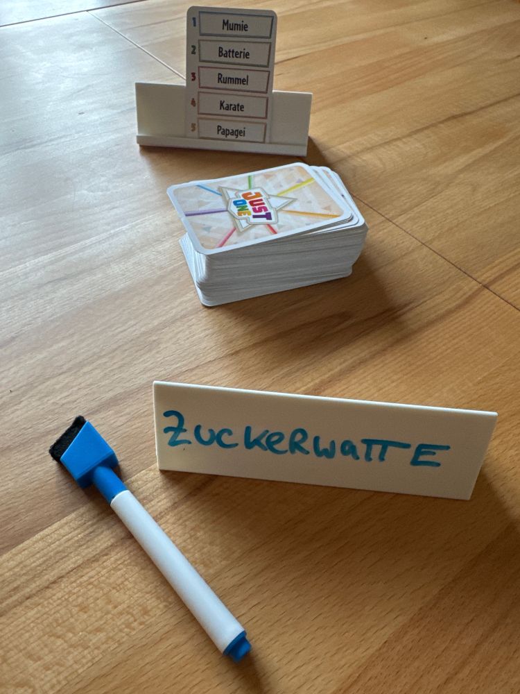 Spielmaterial Just One: Vorgegebene Begriffe auf einer Karte sowie ein geschriebener Begriff