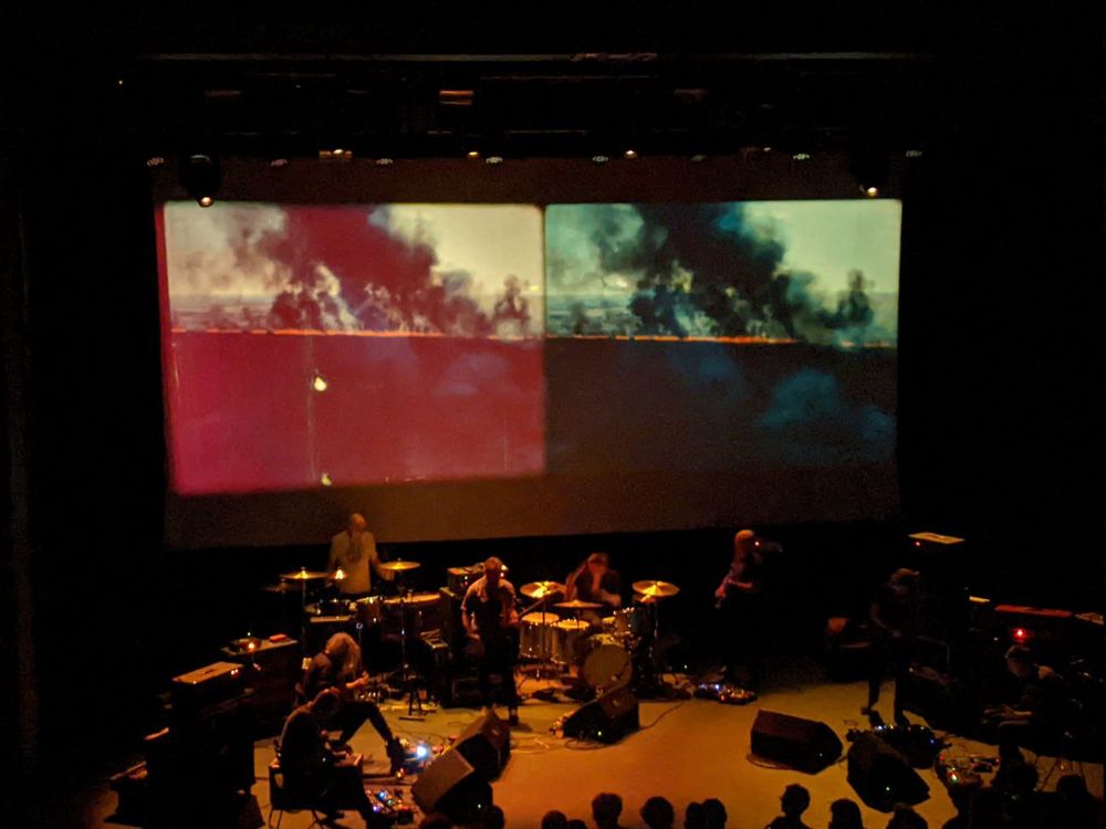 Godspeed You! Black Emperor performing live, under screens showing footage of forest fires.