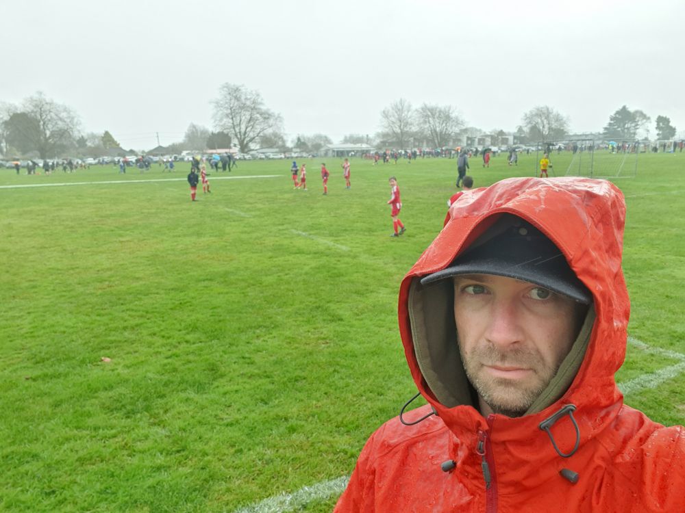 Selfie of the poster wearing a red raincoat in front of a wet children's sports field under a grey, rainy sky