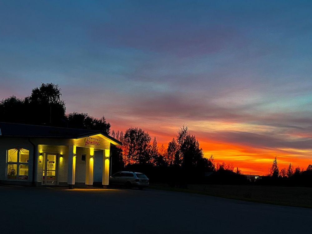 a photo of a bowling alley’s entrance lit up with warm yellow lighting, with the foreground depicting an otherwise dark parking lot and the background depicting a colourful sunset with blue, yellow, orange and the clouds shades of purple