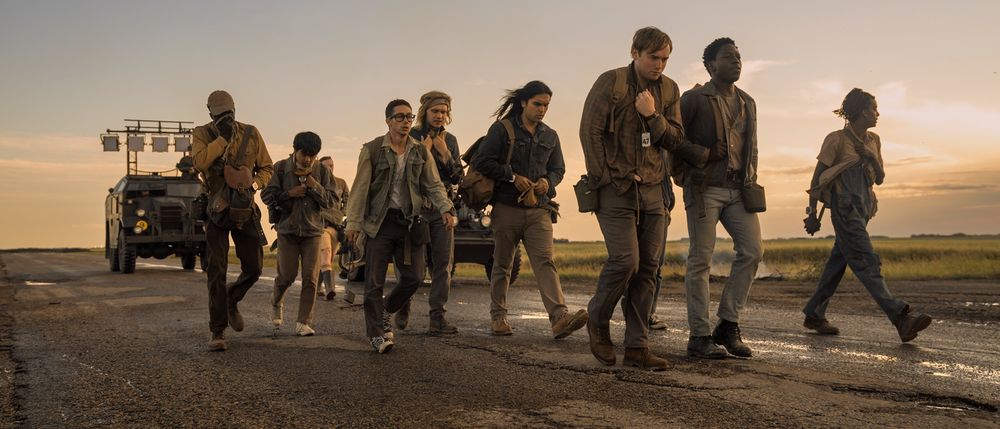 a still photo grabbed from the movie The Long Walk, showing the central characters walking on a tattered asphalt road, with military vehicles following them