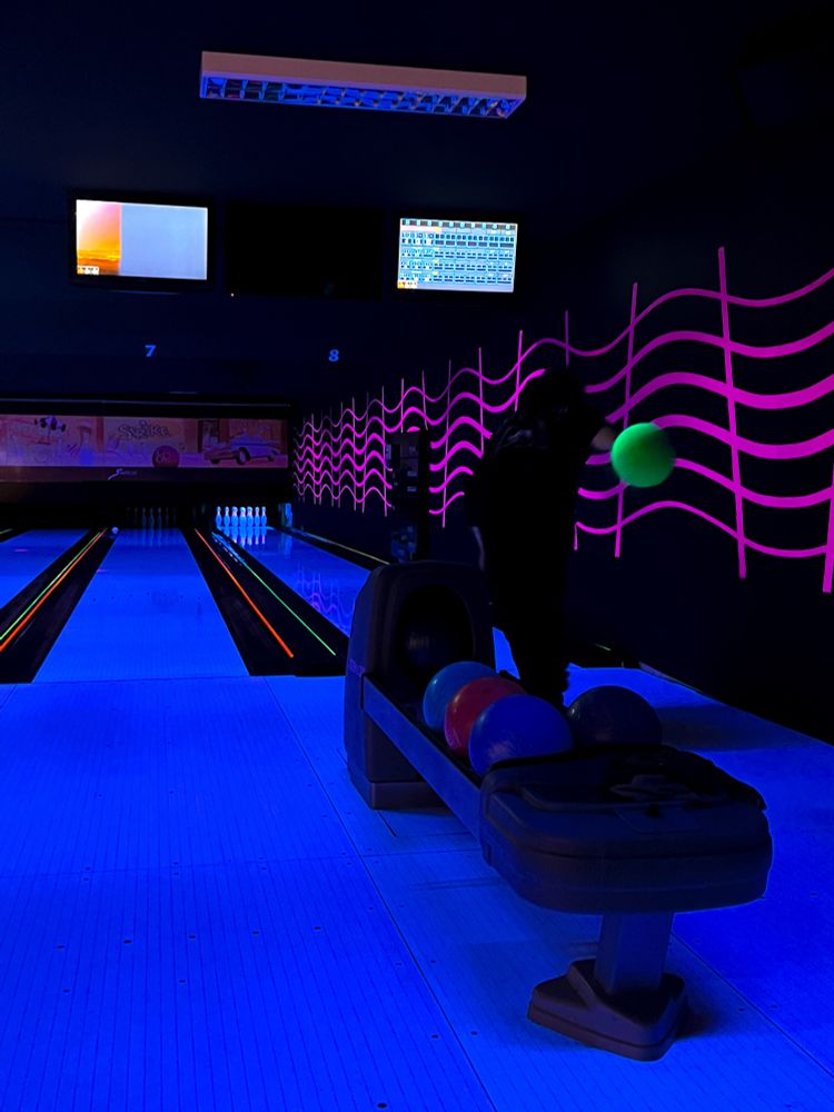 a photo of a person bowling with the alley lit up in blacklight, bringing out neon colours in the decor, the person is mid-throw holding a neon yellow/green bowling ball
