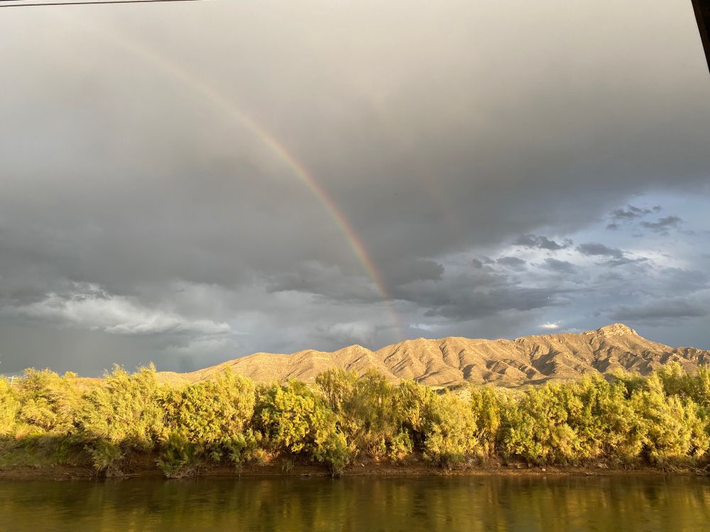 A river in the foreground. Trees and plants are on the other side of the river. A mountain is illuminated behind and above the plants, there’s some mottled gray clouds above that, and a rainbow going from the middle to the top left