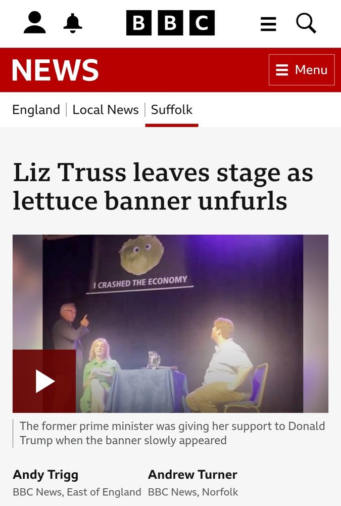 Screenshot of a BBC News webpage, the title of the story is "Liz Truss leaves stage as lettuce banner unfurls", with a photo of Truss, appropriately, in a green dress, on a stage with a banner above her saying "I crashed the economy" under a picture a lecture with googly eyes stuck on it. Under the photo the BBC article sub-heading reads "The former prime minister was giving her support to Donald Trump when the banner slowly appeared", followed by the names of the reporters, Andy Trigg, BBC News East of England and Andrew Turner, BBC News, Norfolk. The lettuce...I mean, Truss, is sitting by a table with a blue table cloth, and two men are with her on stage, one seated at the table looking up at the banner, and the other man behind Truss, standing, pointing up at the banner (like anyone could miss it).