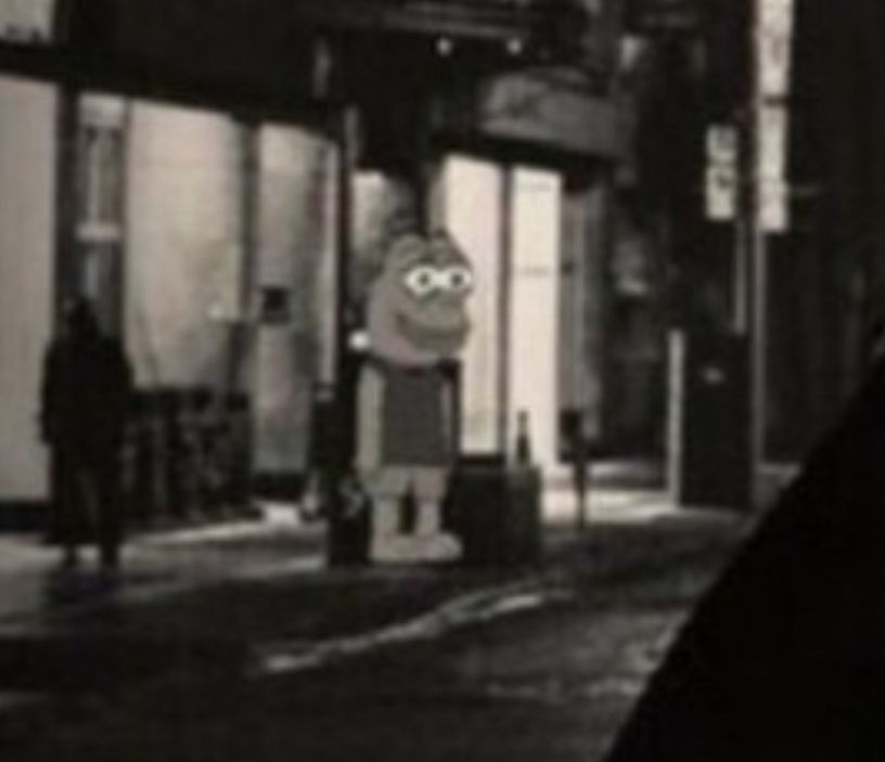 Screenshot of the OP's image, zooming in on the mid-left hand side to show a bug-eyed, fat-lipped cartoon creature wearing a t-shirt and shorts, just standing in the pavement some way behind Trump and looking towards him. WhereTF did they get this image and how did no-one see that??