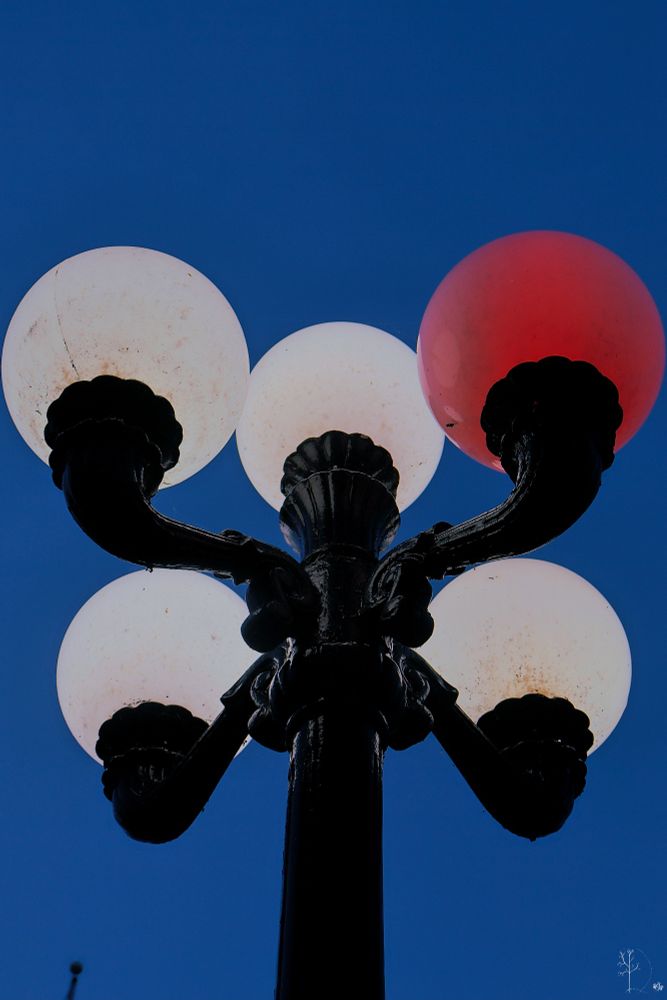 The image shows a black metal street lamp with five spherical bulbs against a deep blue sky. Four of the bulbs emit a white light, while one bulb is distinctly red. The lamp is ornate, with decorative elements at the base of each bulb and curving supports leading to a central pole. The gleaming black metal contrasts sharply with the brightly lit bulbs, creating a striking silhouette effect against the sky.