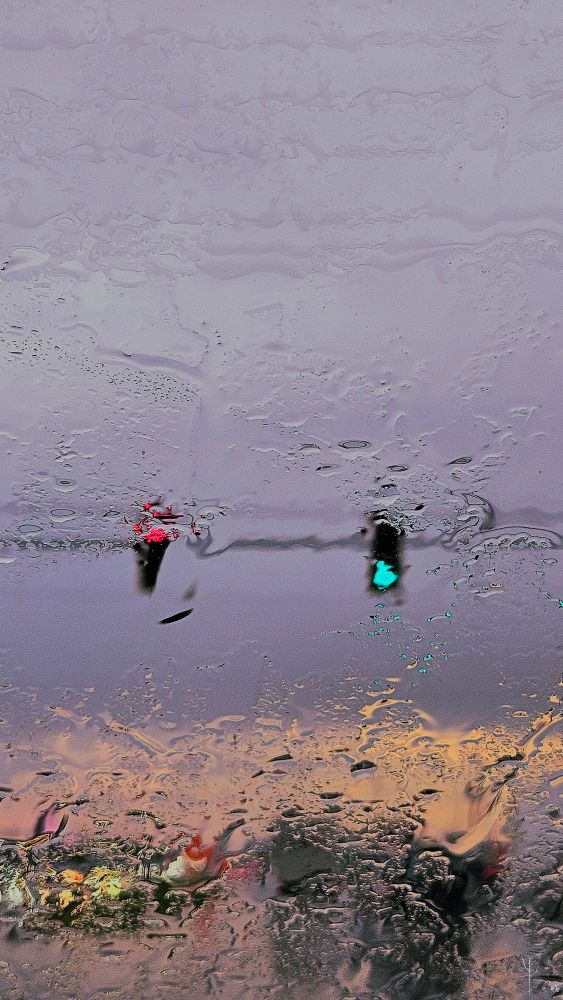 A photograph taken through a rain-soaked window, creating a blurred and distorted view of a city street. Water droplets and streaks on the glass obscure the scene, giving it a dreamy, impressionistic quality. In the upper part of the image, two traffic lights glow through the rain—one red on the left and one green on the right—suspended against a muted, cloudy sky. Below, the faint outlines of cars and headlights shimmer in the wet reflections, blending with warm tones of sunset to produce a painterly mix of purples, oranges, and dark hues.