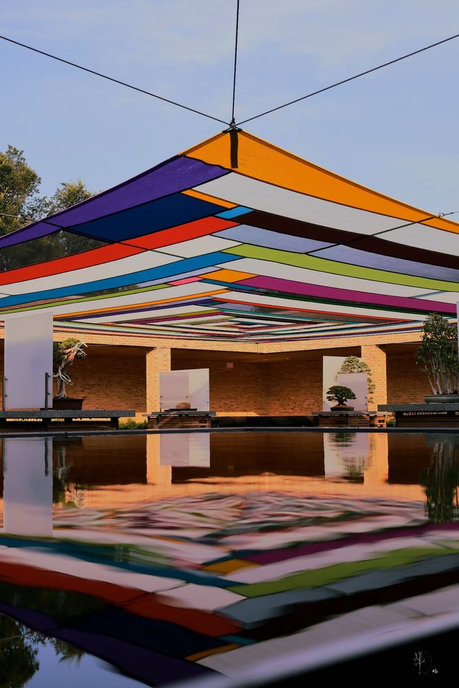 A vibrant overhead canopy made of multicolored fabric panels stretches across a courtyard, suspended by cables from four corners. The geometric arrangement of the panels features bold shades of orange, purple, blue, green, red, and white. Below, a still reflective pool mirrors the colorful canopy and the surrounding space. The courtyard is framed by warm-toned brick walls and features bonsai trees displayed on platforms, contributing to a serene and artistic atmosphere. The sky is lightly clouded, allowing soft natural light to illuminate the scene.