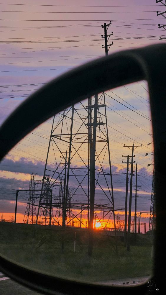 The image captures a sunset scene viewed from a car's side mirror. The foreground is dominated by a silhouette of a transmission tower and several utility poles, with power lines stretching across the scene. The horizon is colored in warm shades of orange and pink, while the sky above transitions to a soft lavender and blue, suggesting the evening time. In the bottom left corner, part of the car's mirror frame is visible, adding a framing effect to the composition.