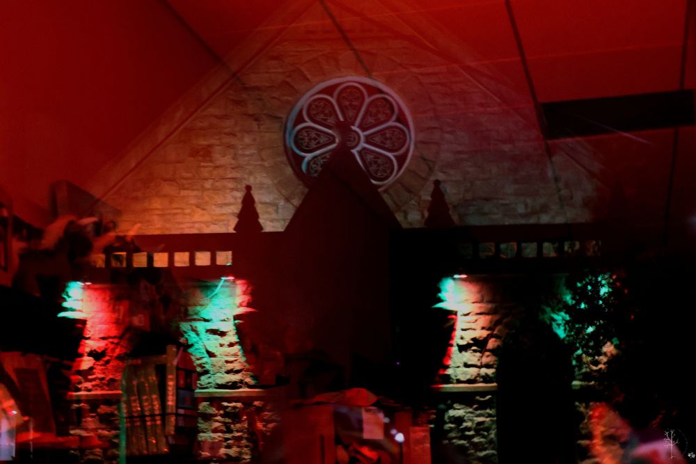 A dim, atmospheric scene showing the reflection of a stone wall with an ornate circular window, cast onto a dark interior space. Red and green lights illuminate sections of the stone texture and surrounding objects, creating layered, ghostlike overlaps. The triangular roofline and silhouetted architectural details appear doubled due to the reflective surface.