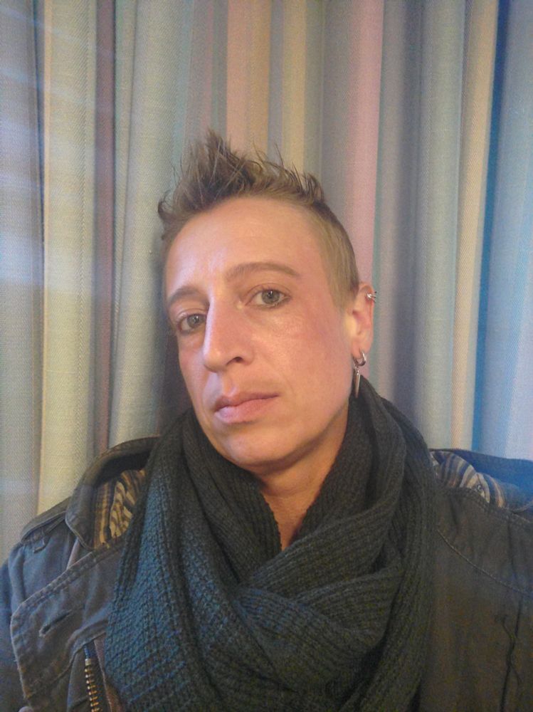 Ein Selfie mit ernstem Blick. Meine Haare sind stachelig aufgestellt, ich trage rechts einen hängenden Stachel Ohrring und einen Helix Clip in Silber. Ich habe meinem Flaum zu einem Schnauzbart eingefärbt. Ich trage einen olive farbenen Schaal und einen Parker in dunkel Olive. 