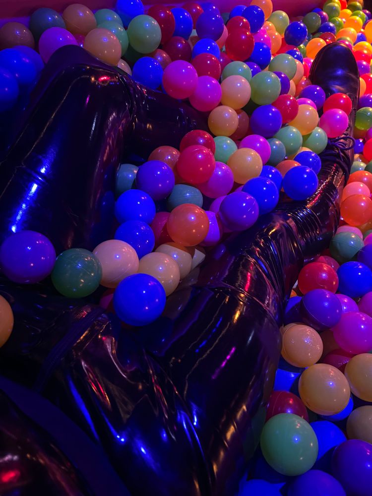 POV photograph showing the waist down of a person in a latex catsuit laying in a ballpit