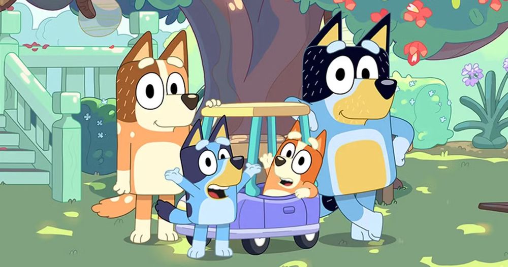 A cartoon of a family of Australian cattle dogs, both blue and red. One red pup sits in a play car, another blue pup alongside with their arms up. The parents (one red, one blue) stand on either side, resting on the play car. Behind is a large tree (only the trunk and lower canopy visible), other bushes visible and wooden steps and low balcony, presumably a house.