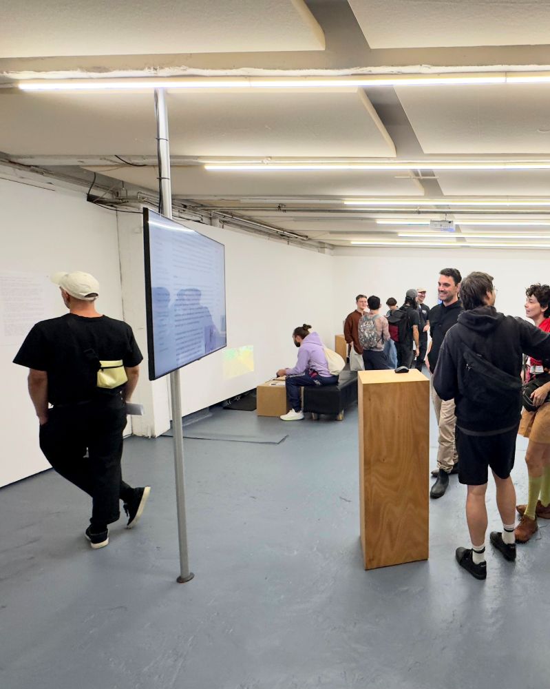 A group of people inside a white-walled gallery space with fluorescent ceiling lights. A flat screen is mounted on a pole showing text, while visitors walk around or sit on black benches. Some are standing in conversation, while one person in a black shirt and cap looks at the screen.