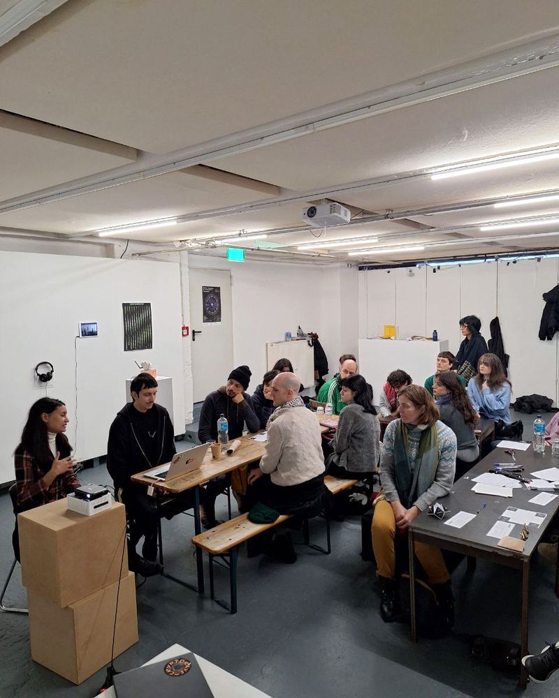 A woman sits at the front of a room speaking to a group gathered around a long wooden table. Participants listen attentively, some taking notes or using laptops. The setting is an art workshop space with white walls and a ceiling-mounted projector.