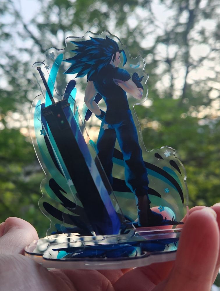 An acrylic standee of Zack Fair from Final Fantasy 7, held out in front of a blurry background of leaves.  The standee features Zack facing away wistfully and holding a hand out to catch a single white feather, with the Buster Sword standing in the ground next to him.