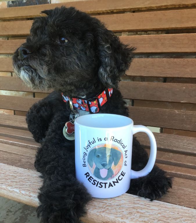 Henry a rescue black and white rescue cockapoo, lays on a wooden bench with a coffee mug from Lissa Grey that has a black dog on it and says "Being joyful is a radical act of resistence" 